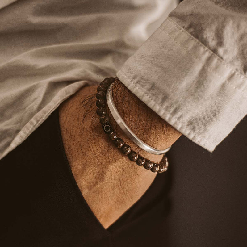 Een man in een wit overhemd draagt de bruine Ninetwofive Albuna kralenarmband voor heren en een zilveren Cuff, met zijn hand in zijn zwarte broekzak.