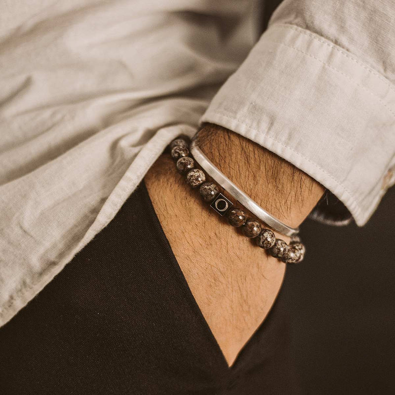 A man in white shirt wearing the Ninetwofive Albuna brown men's beaded bracelet and a cuff, with his hand in his black trouser pocket.