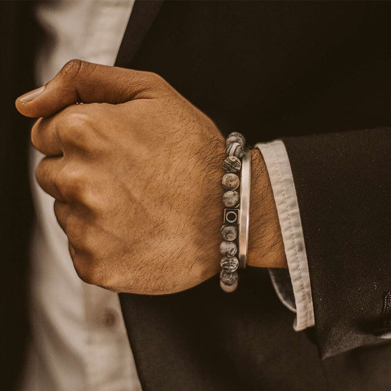 Een man in een zwart-wit pak draagt een grijze marmeren kralenarmband van Alrazas en een heren-Cuff van Ninetwofive.