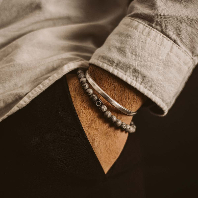 Ein Mann in einem weißen Hemd, der das graue Perlenarmband „Alrazas“ für Herren von Ninetwofive sowie ein silbernes Armband trägt und die Hand in die Hosentasche seiner schwarzen Hose gesteckt hat.
