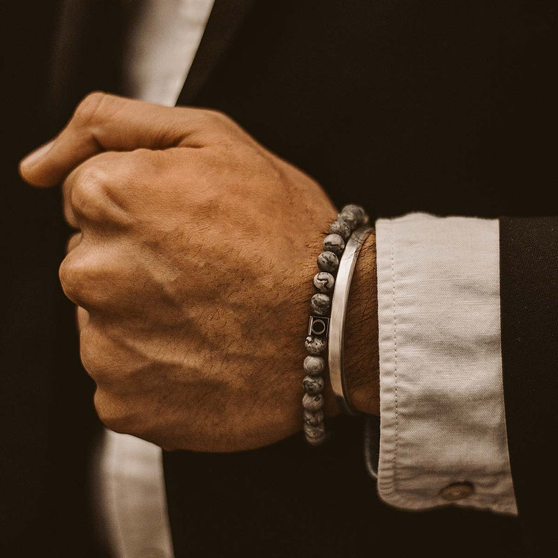 Ein Mann in einem schwarzen Anzug trägt ein graues Marmorperlenarmband von Alrazas und ein Herrenarmband von Ninetwofive.