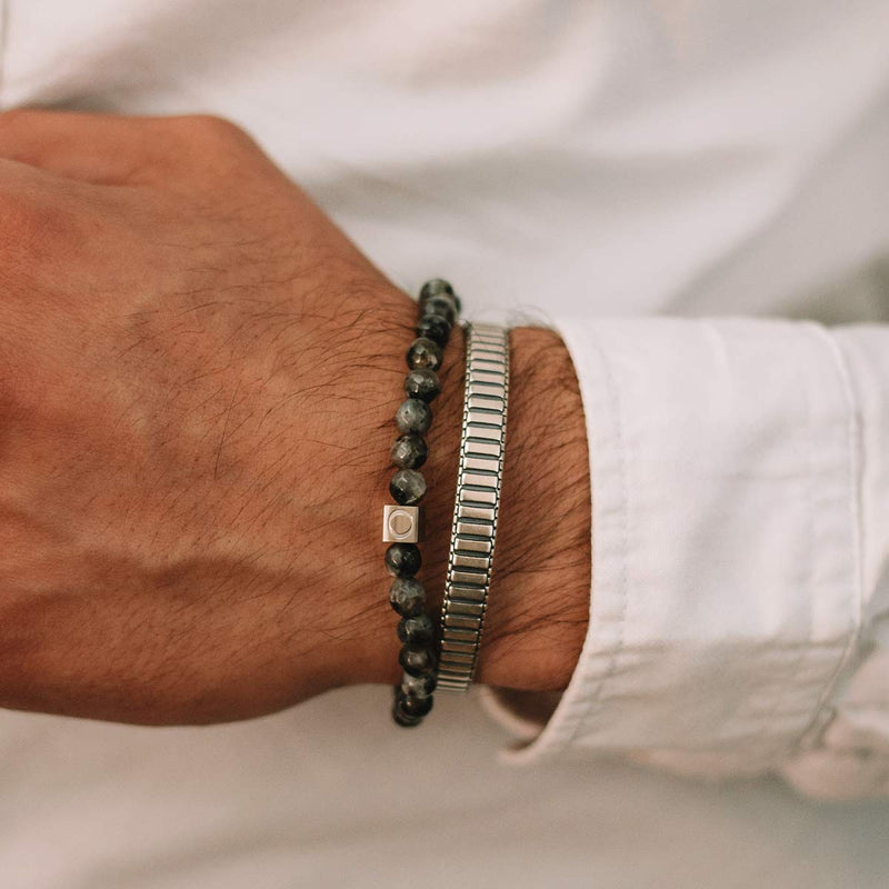 A man in a white long sleeve shirt is wearing the Aswad mens black beaded bracelet adorned with a spectrolite stone and a silver cuff bangle by Ninetwofive.