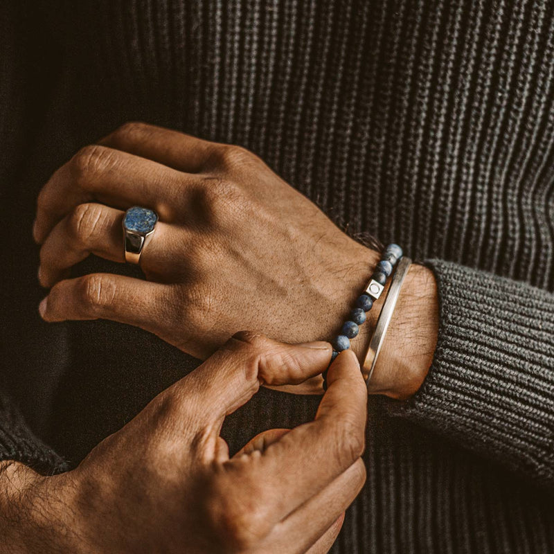 Man in een zwarte trui met een armband van Azraq-sodaliet, een zilveren arm bangle en een heren horloge zegelring van Ninetwofive.