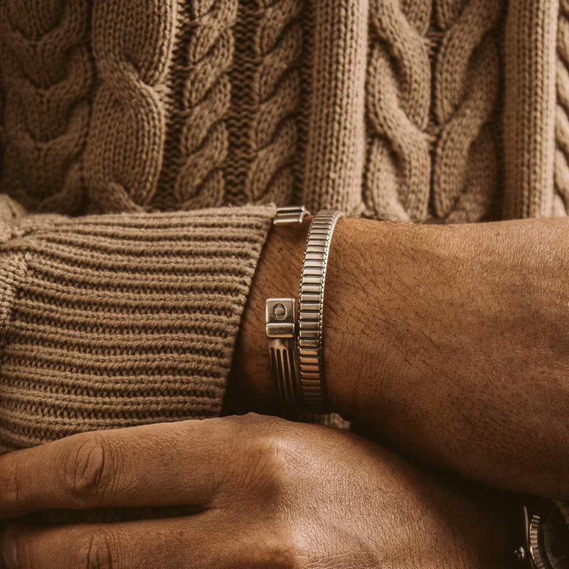 Ein Mann, der den Kenan-Armreif aus Sterlingsilber für Herren (7 mm) sowie ein Armband zu seinem braunen Pullover trägt.