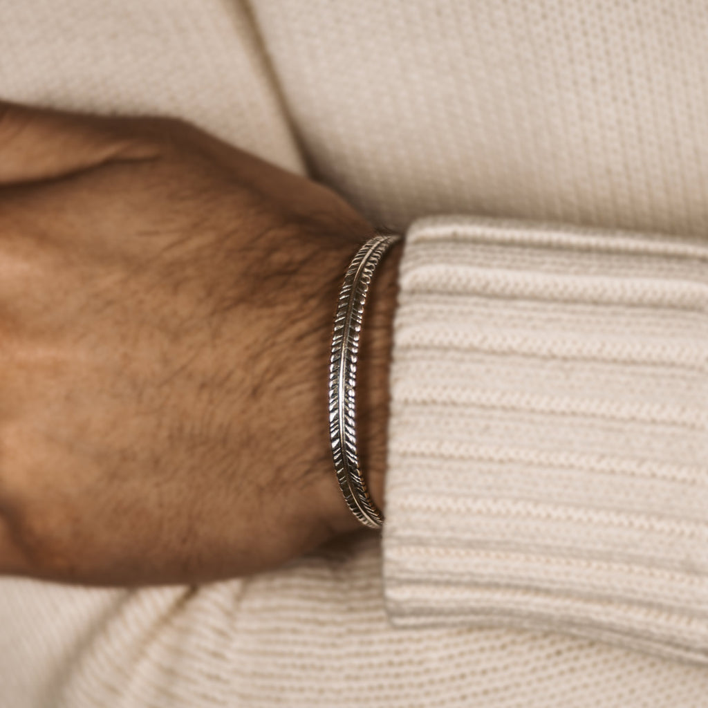 A man wearing a white sweater and a Zahir - Thin Sterling Silver Feather Bangle 6mm.