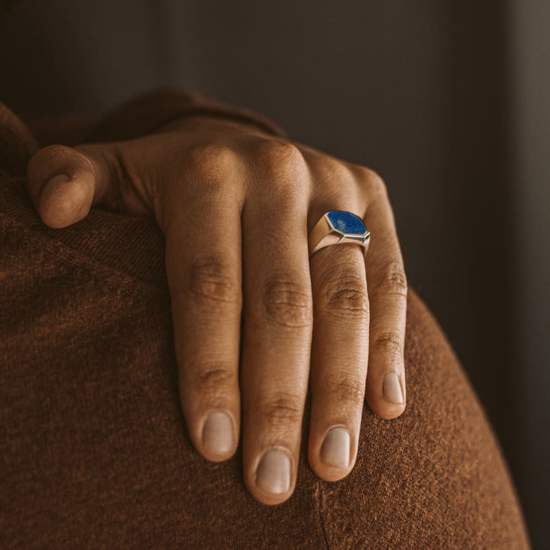 Man in a brown sweater wears the Kadar silver ring, placing his hand onto his shoulder to show the blue stone.