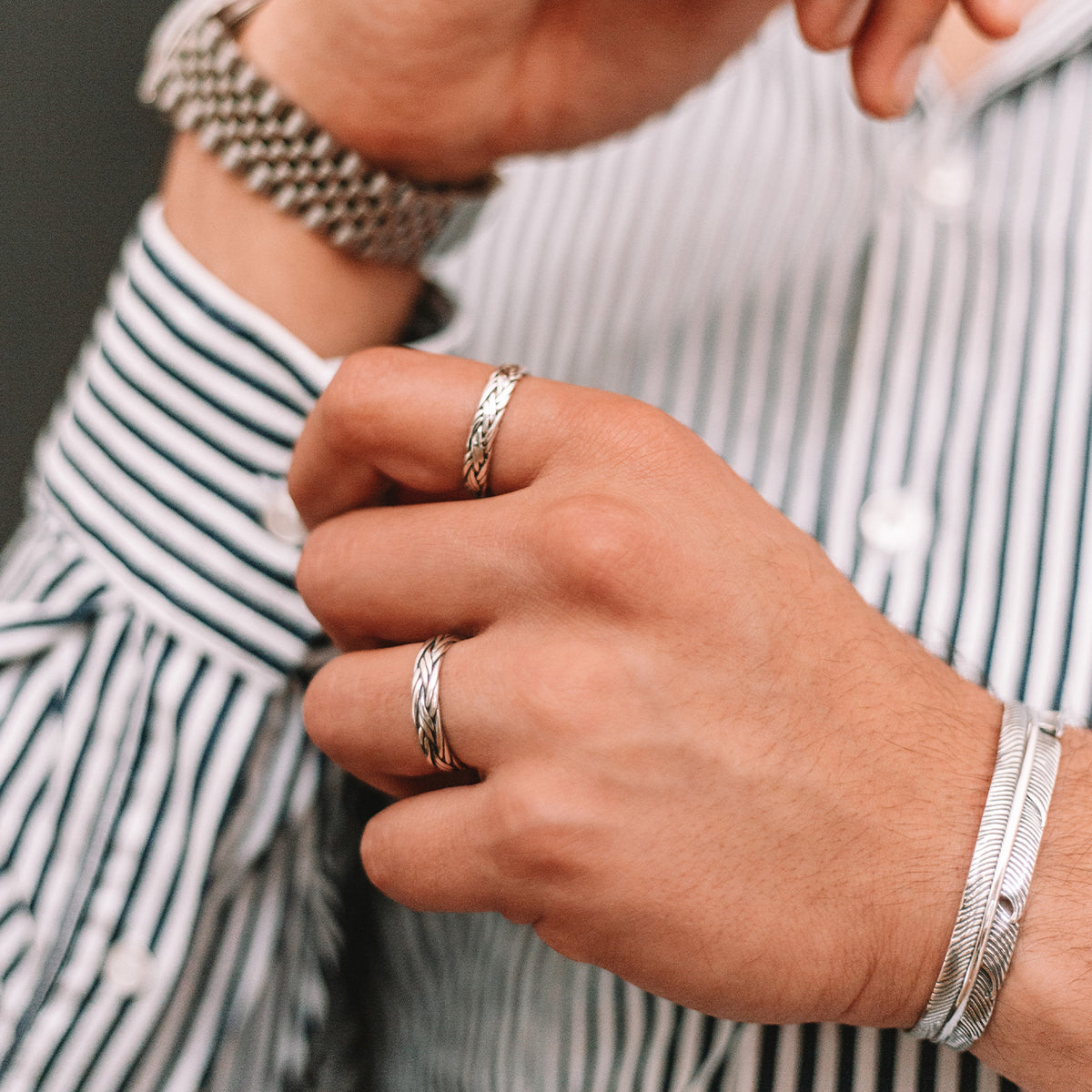 Een man met een gestreept overhemd en zilveren ringen, waaronder een gegraveerde Latif - Dunne Sterling Zilveren Gevlochten Ring van 5 mm.