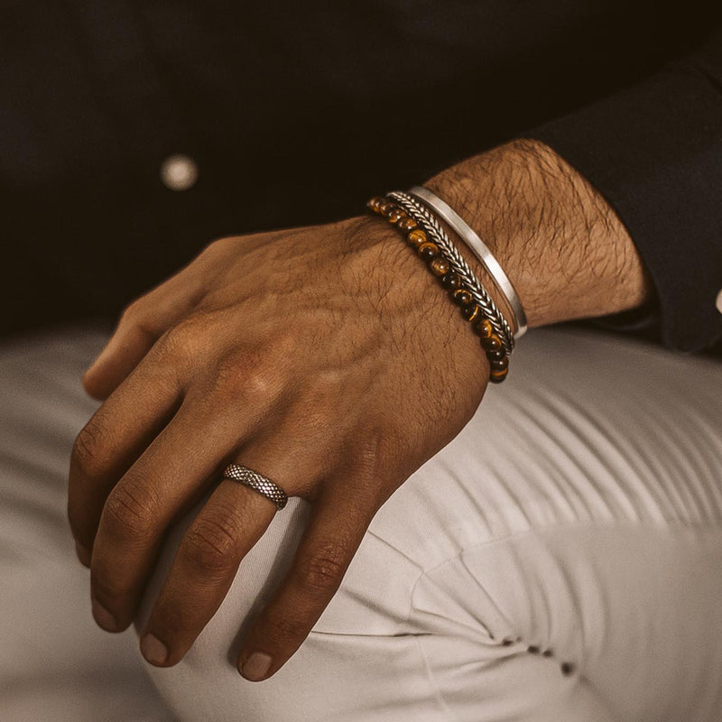 Een man die een zilveren Ninetwofive-Cuff, een armband met tijgeroogkralen en een zilveren ring aan zijn ringvinger draagt.