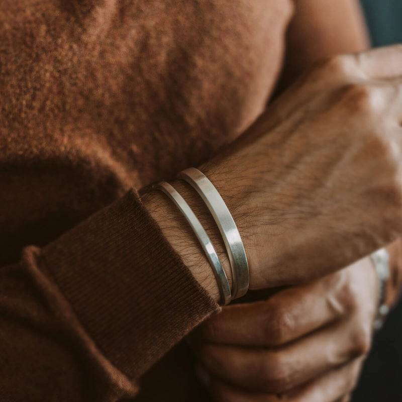 Een man in een knusse bruine trui die de sterlingzilveren cuff heren armbanden van Noor draagt, van Ninetwofive.
