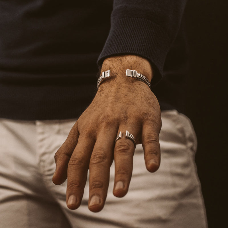 Ein Mann in schwarzem Pullover und weißer Hose zeigt seine Hand mit dem Mateen oxidierten Silber-Bangle und einem Herrenring, Ninetwofive.
