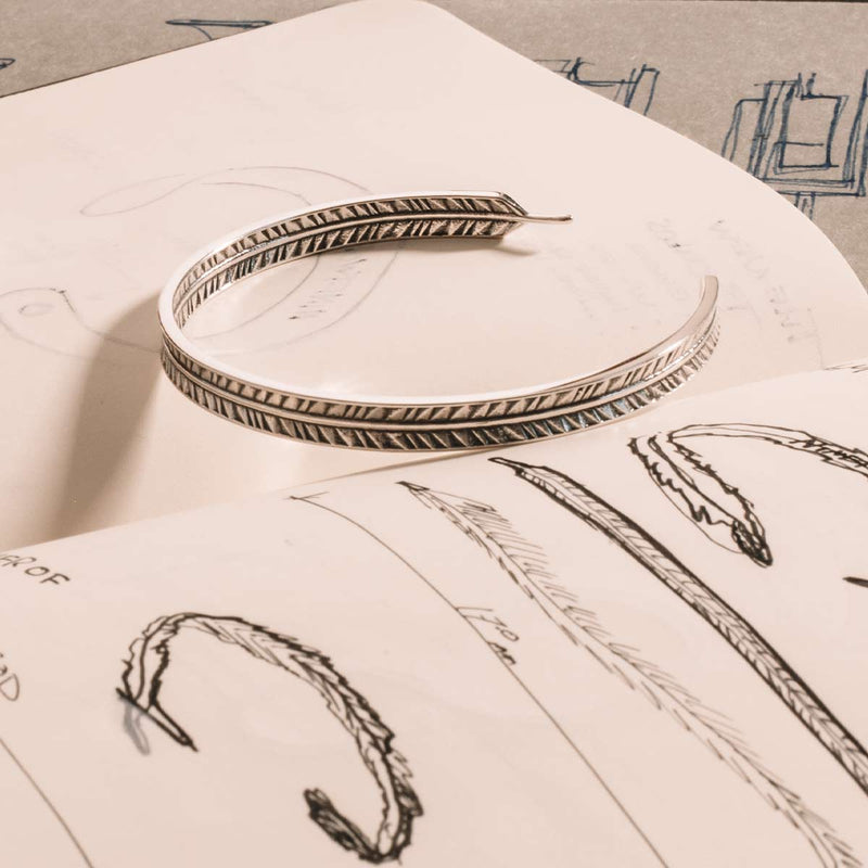 Le bracelet pour homme « Zahir thin » en argent 925, orné d'une plume, est présenté posé sur un carnet de croquis.