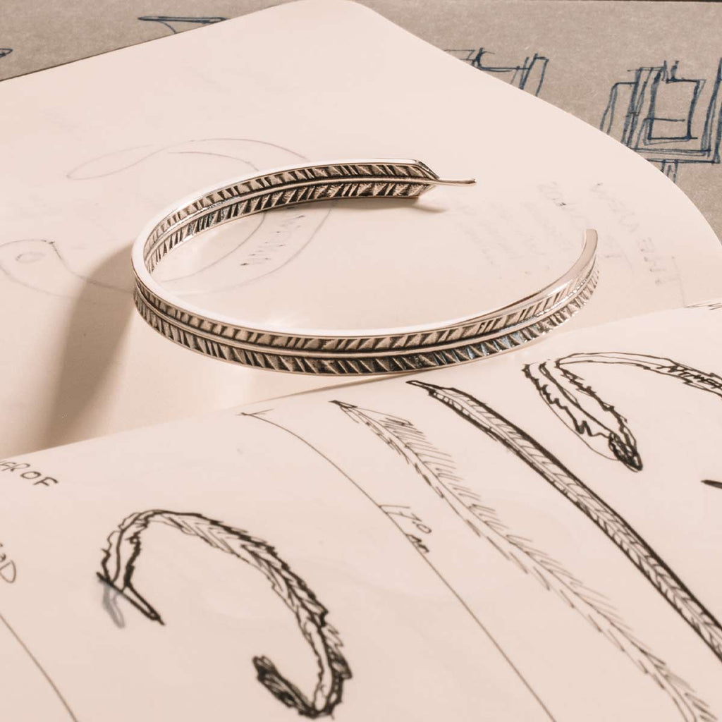 A Zahir - Thin Sterling Silver Feather Bangle 6mm atop a book.