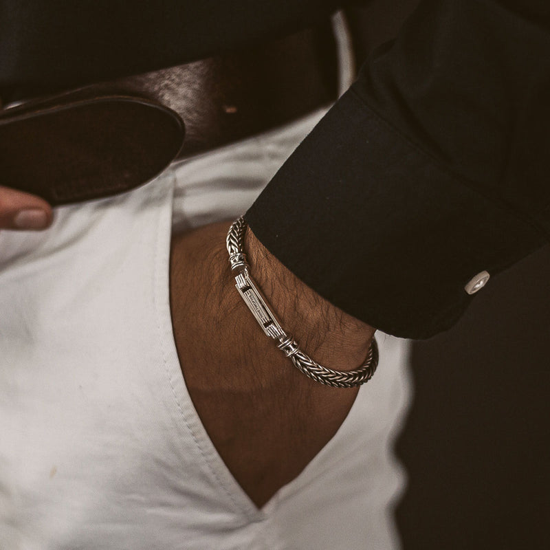Een man in een zwart overhemd en een witte broek met een bruine riem draagt de zilveren gevlochten Adam-armband voor heren, terwijl hij zijn hand in zijn zak laat rusten, Ninetwofive.