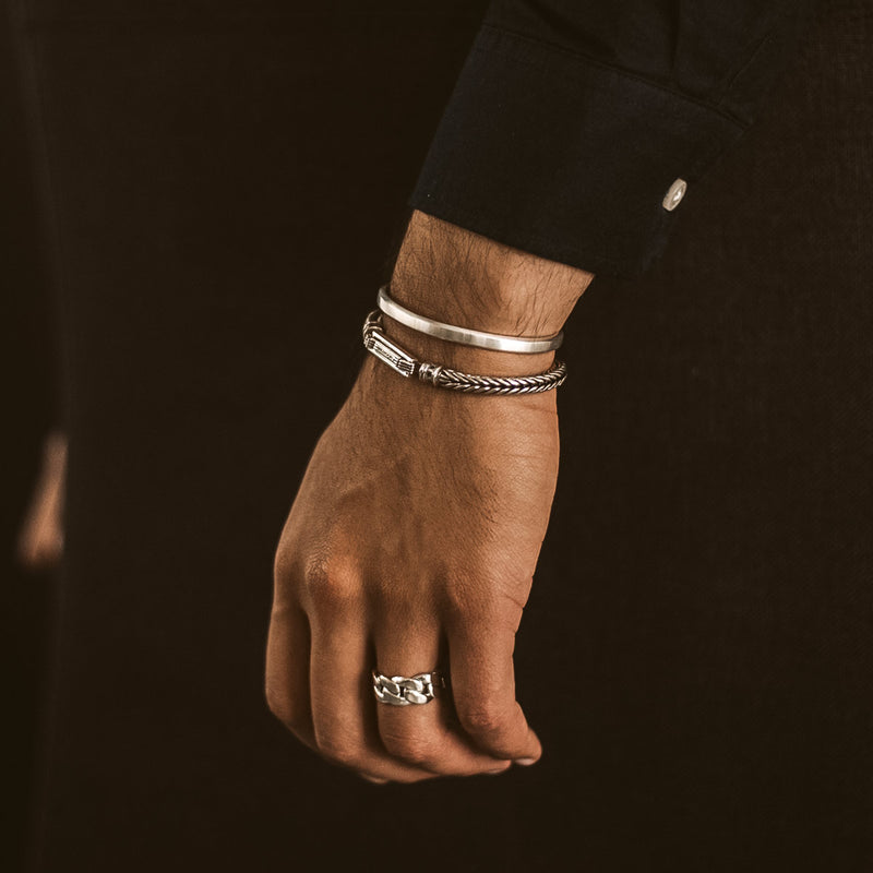 Een man in een zwart shirt met lange mouwen, die de zilveren gevlochten armband, ring en manchet van Adam voor heren draagt, afkomstig van Ninetwofive.