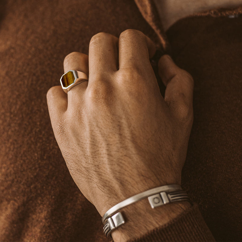 De hand van een man met de Ninetwofive Alem Tijgeroog Zegelring aan zijn ringvinger, gecombineerd met een heren armband en zilveren cuff.