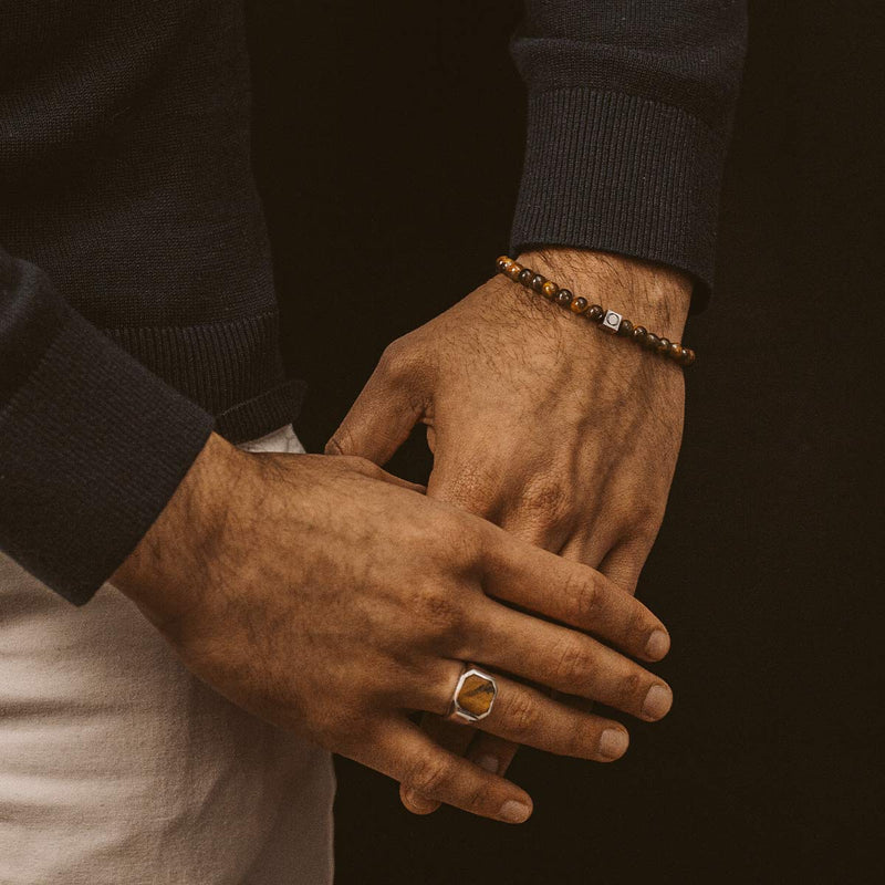 Een man laat een Alnamr-armband voor heren met tijgeroogkralen en een zilveren zegelring zien, afkomstig van Ninetwofive.